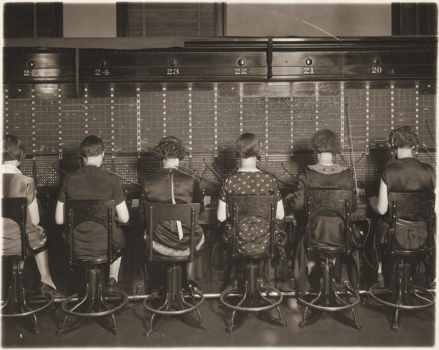 Six telephone operators seen from behind, seated in a row at a switchboard of numbered positions, 1927.