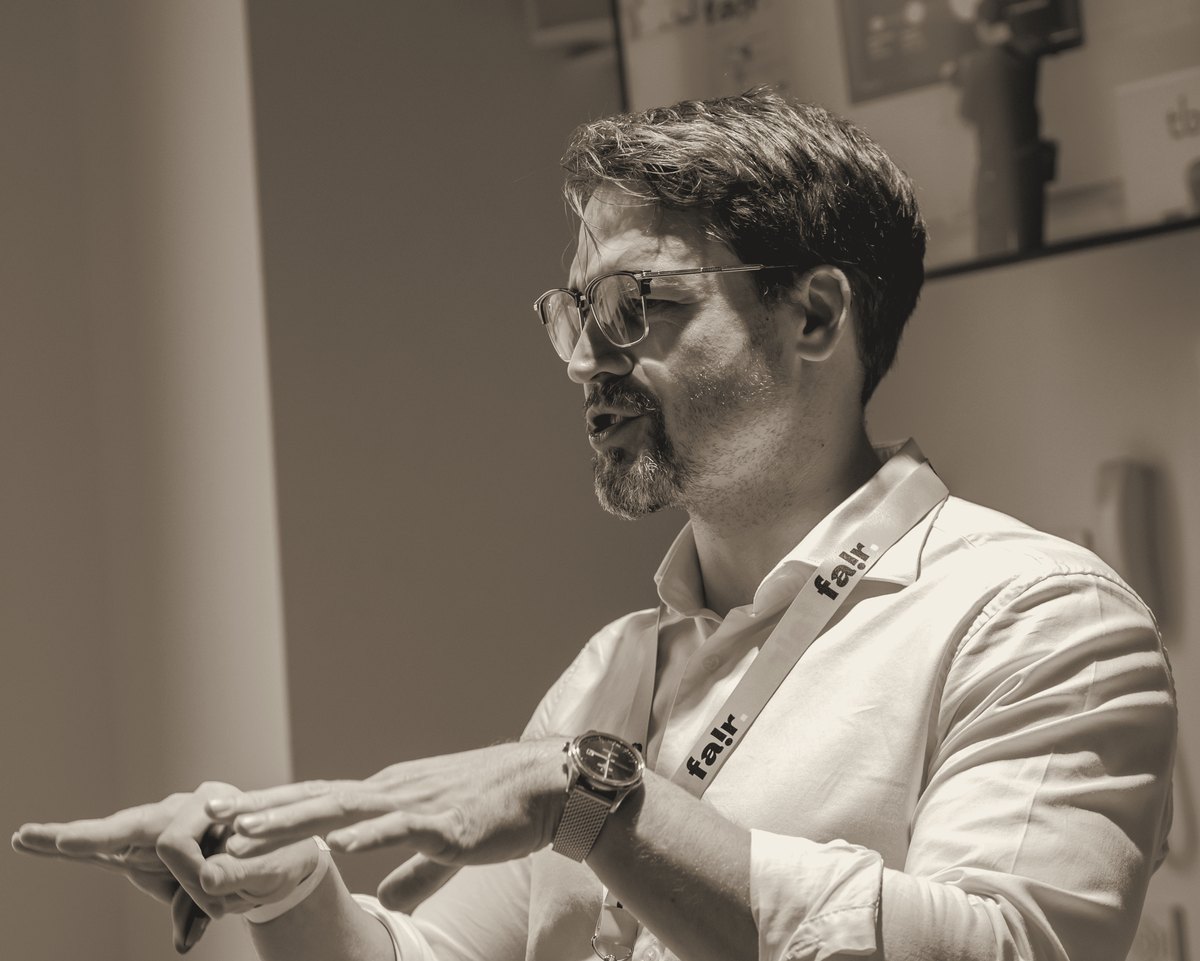 Ivan Nosov explaining with both hands held level, glasses, white shirt, FAIR lanyard.